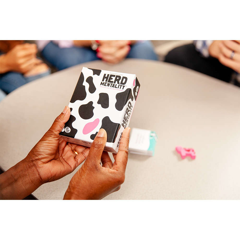 Hands holding the cow-spotted box for the "Herd Mentality" party game, played by a group