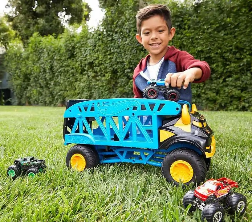 A smiling boy plays with a large blue toy monster truck carrier and small monster trucks on the grass