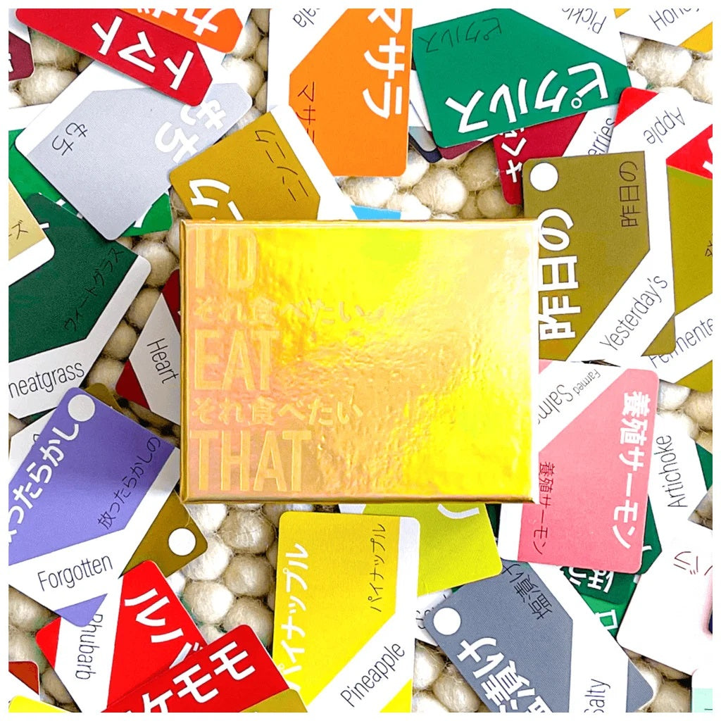 A golden holographic I'd Eat That Card Game box is surrounded by scattered colourful cards with English and Japanese words like pineapple and artichoke, on a textured white background