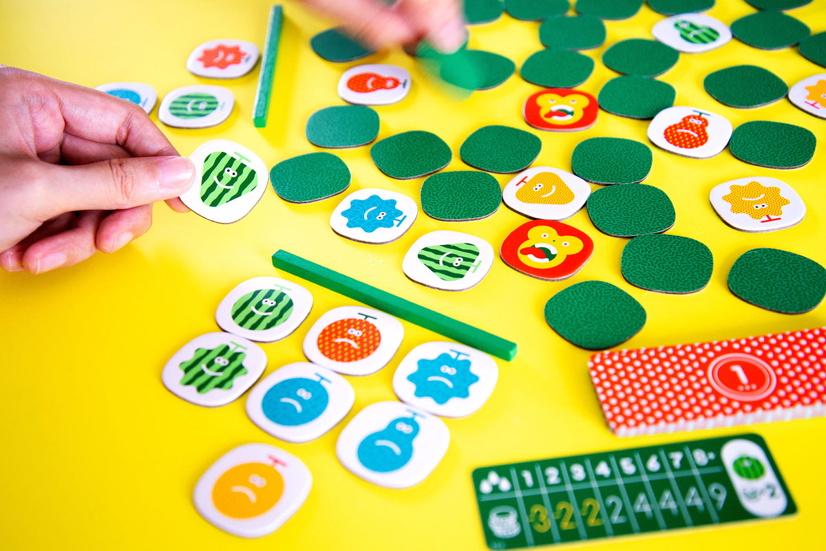 A hand places a happy watermelon game token on a bright yellow game board with other colourful fruit pieces