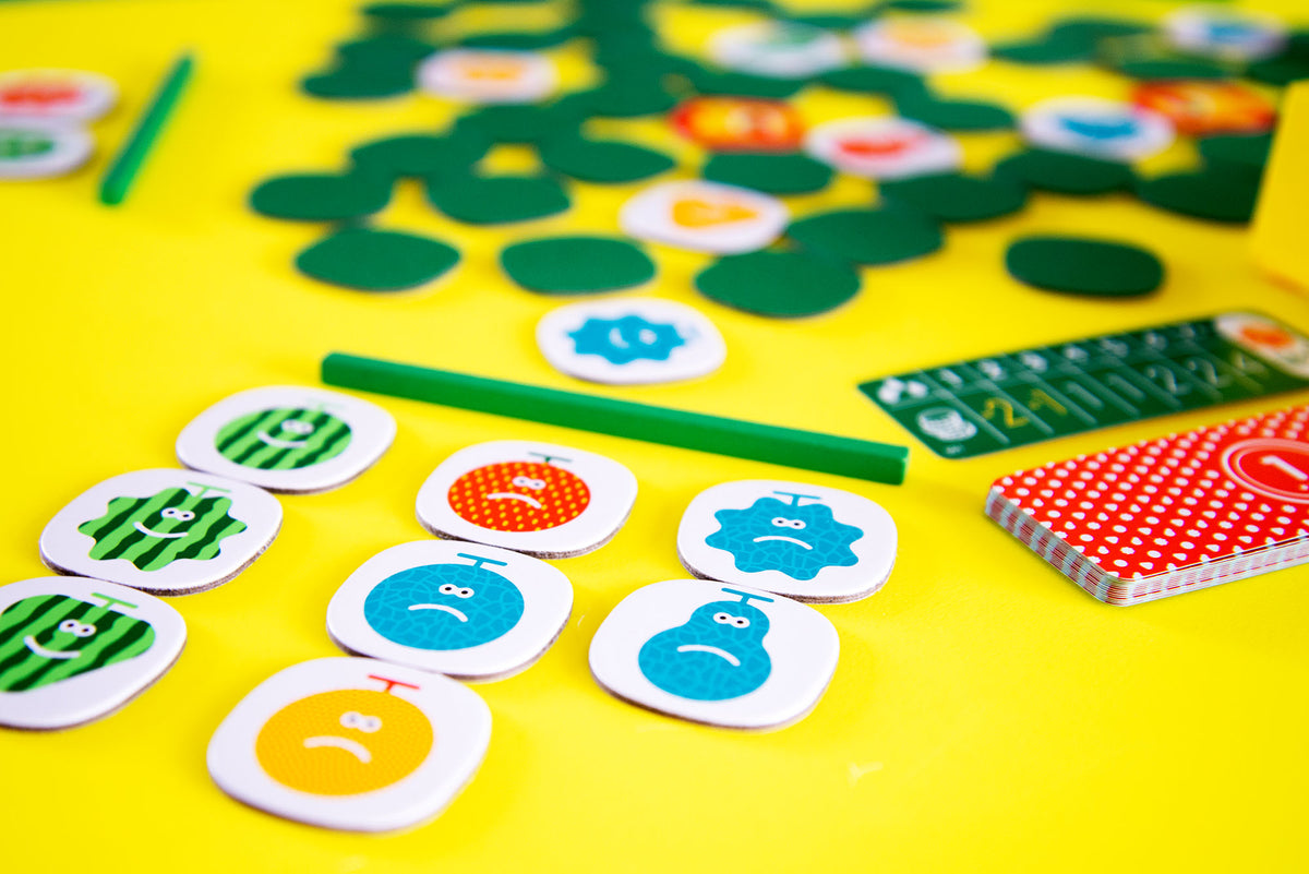 Colourful board game pieces including happy watermelon and sad fruit character tokens green leaf shapes cards and a score tracker laid out on a bright yellow surface