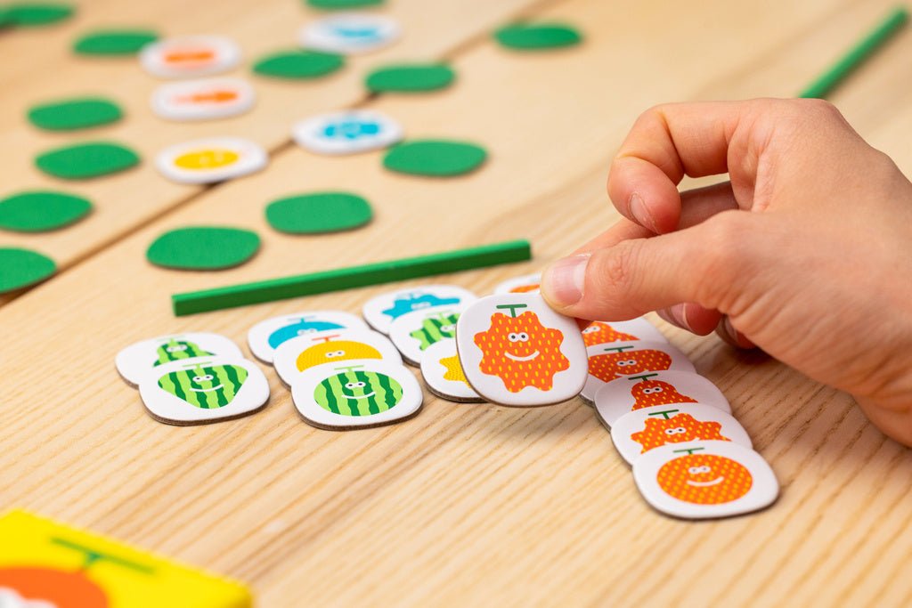 A hand selects a smiling orange fruit game piece from a stack on a wooden game board with other colourful tokens