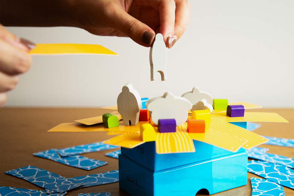 Person playing Oink Games Rafter Five balance puzzle placing a white wooden human figure onto a blue box with yellow patterned boards and colorful cubes