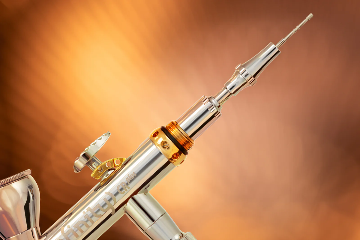 A detailed close-up of a shiny silver and gold &quot;Infinity CR plus&quot; airbrush, angled for precision painting
