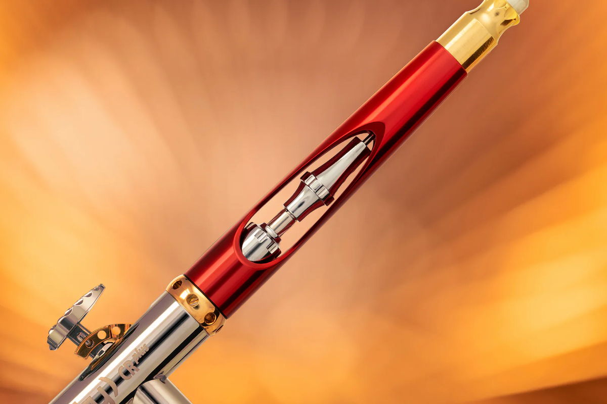 A sleek red, silver, and gold airbrush with its internal mechanism visible through a slotted barrel