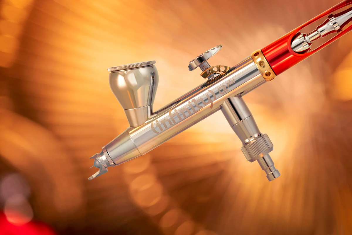 A silver and red &#39;Infinity CRplus&#39; airbrush with a top-feed cup and gold accents, against a warm, blurry orange background