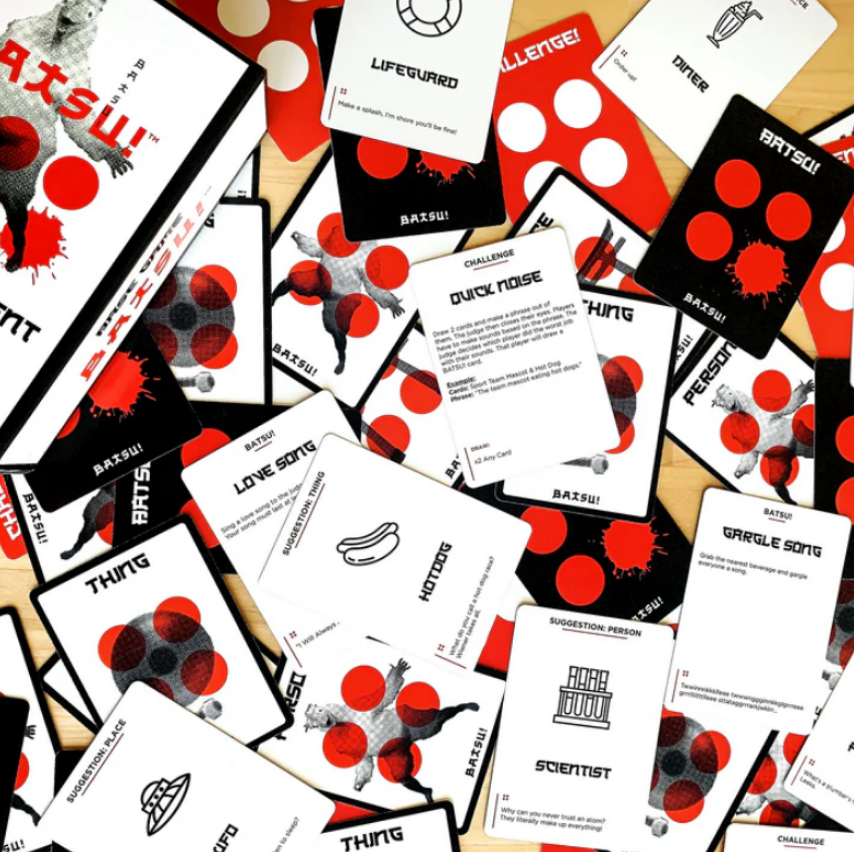 Batsu! game box and many cards scattered on a surface. Cards are black/white with red splatters, text, and silhouettes