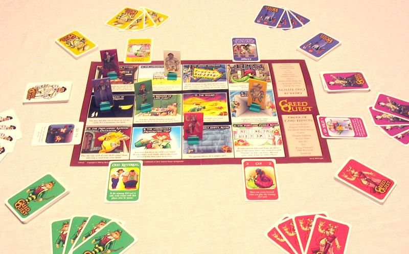 The Greed Quest board game is laid out on a table, featuring a central game board, character pieces, and various piles of colorful cards