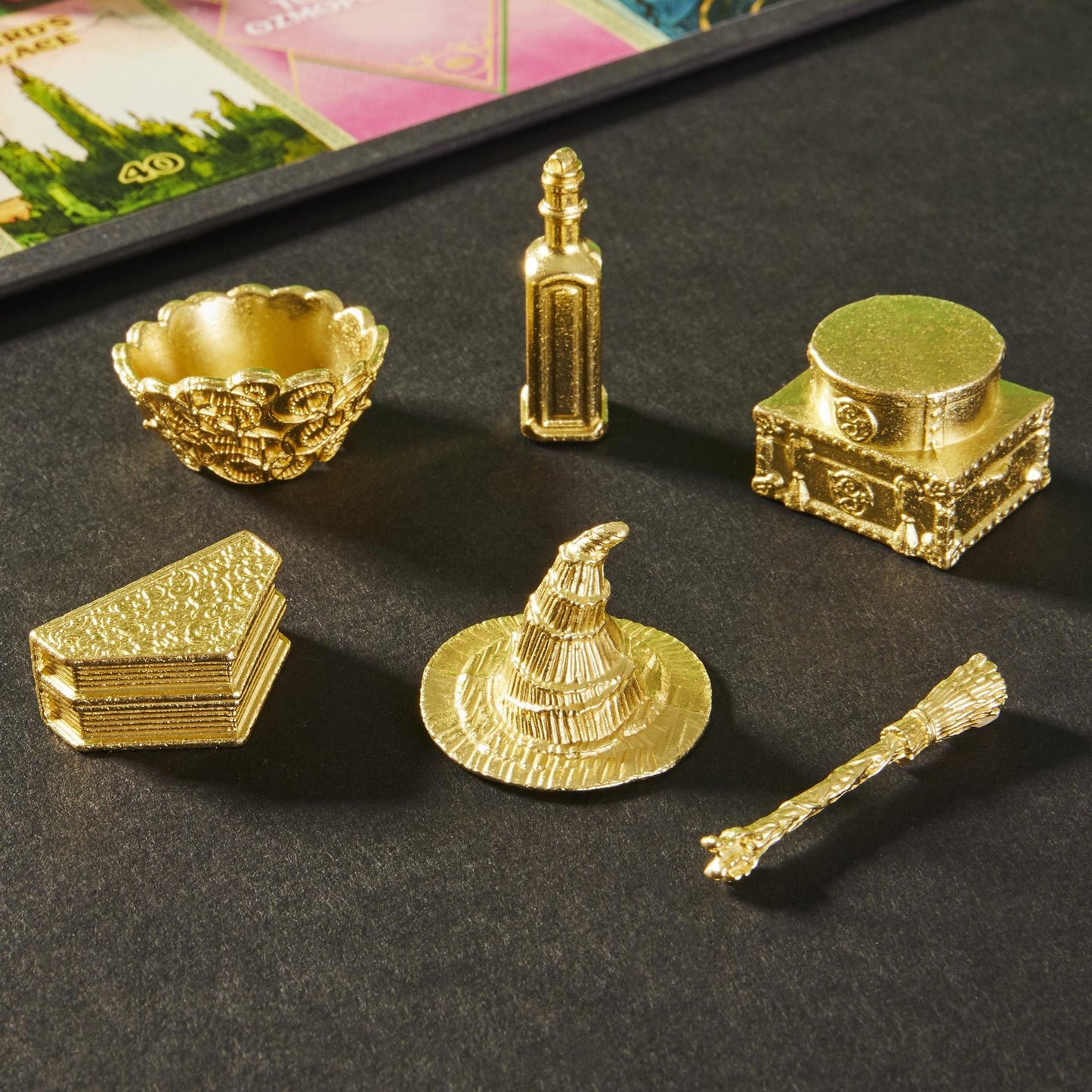 Six ornate gold fantasy game tokens: a cauldron, bottle, chest, spellbooks, witch's hat, and broom, on a dark surface