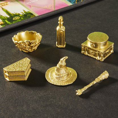 Six ornate gold fantasy game tokens: a cauldron, bottle, chest, spellbooks, witch's hat, and broom, on a dark surface