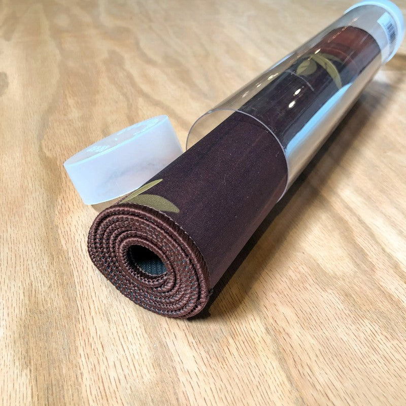 A rolled dark brown desk mat with a pattern, partially emerging from its clear plastic tube on a wooden surface