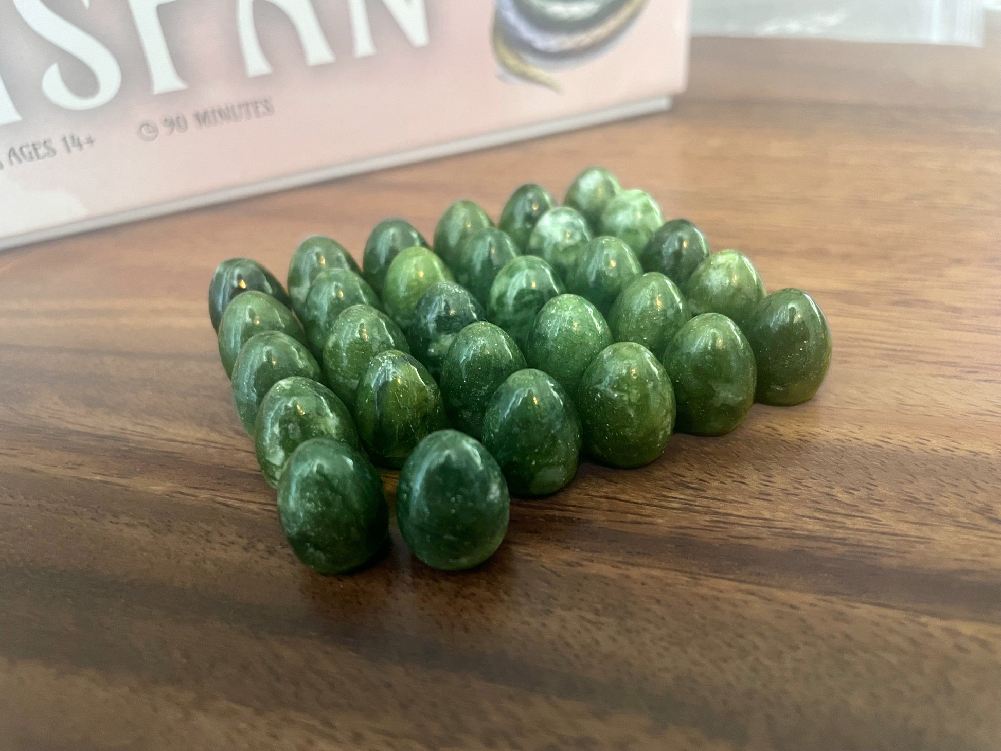 Polished green, egg-shaped game tokens clustered on a wooden table, near a blurred board game box