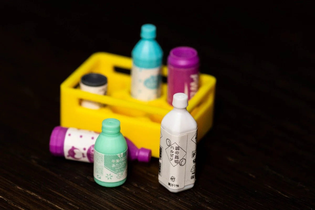 A collection of colourful miniature bottles and containers, some in a yellow crate and others on a dark wooden surface, resembling Tokyo Jidohanbaiki vending machine items