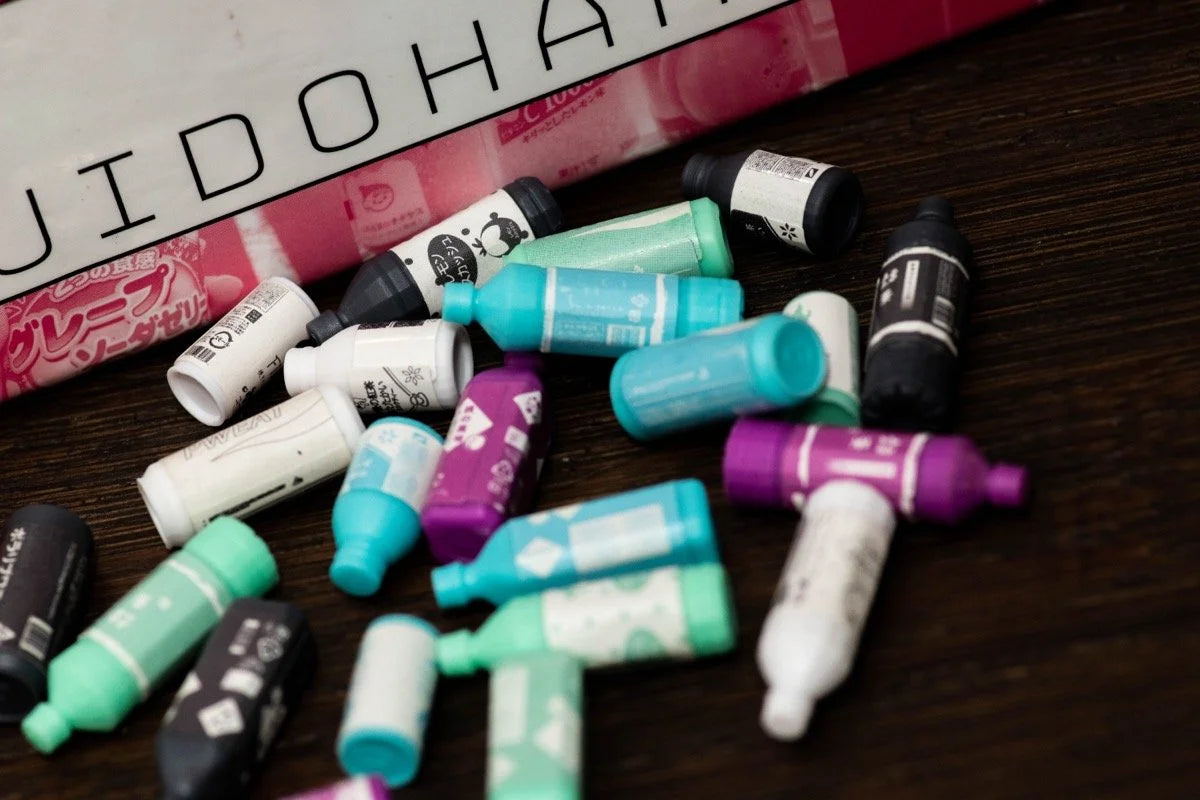 Many colourful Tokyo Jidohanbaiki miniature bottles are scattered on a dark wooden surface, with a white and pink background showing text