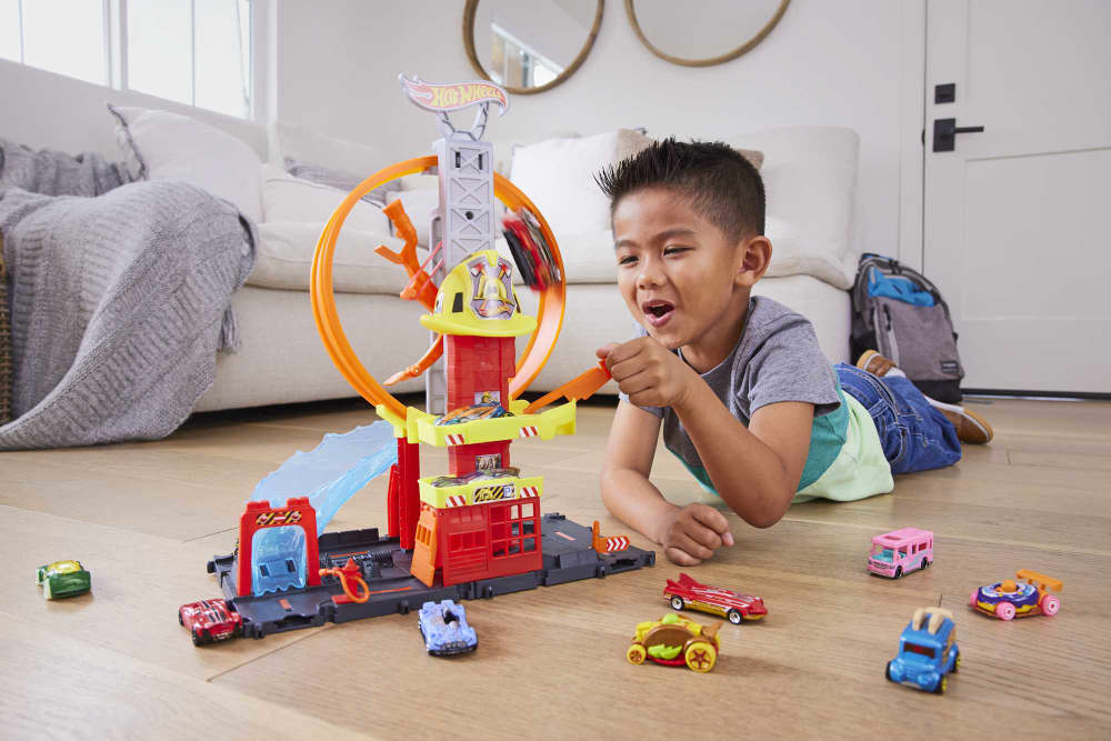 Excited boy plays with Hot Wheels track set, launching a car on its loop; toy cars on the floor