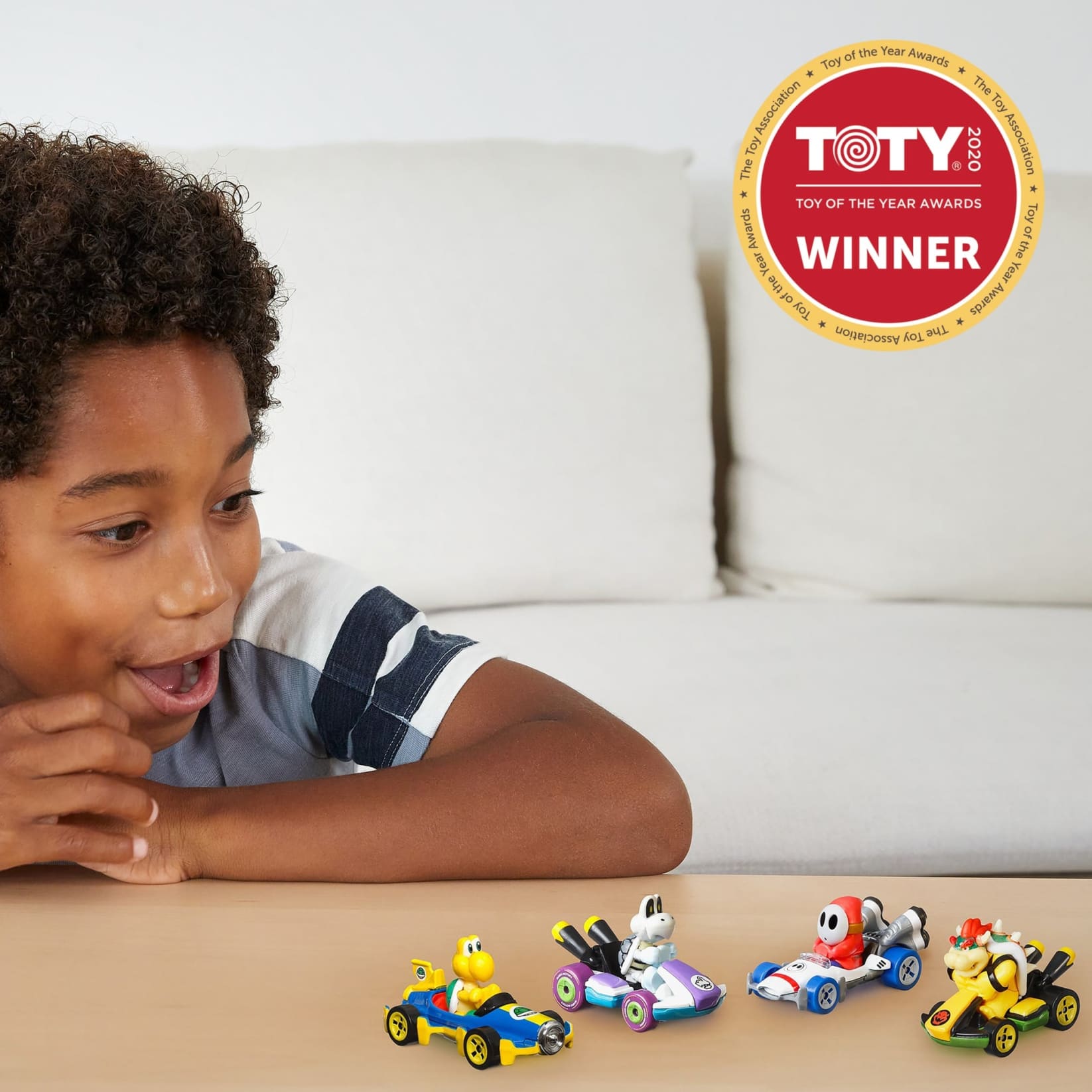 Boy reacts excitedly to four Mario Kart toy cars on a table. A "TOTY 2020 Winner" badge is visible