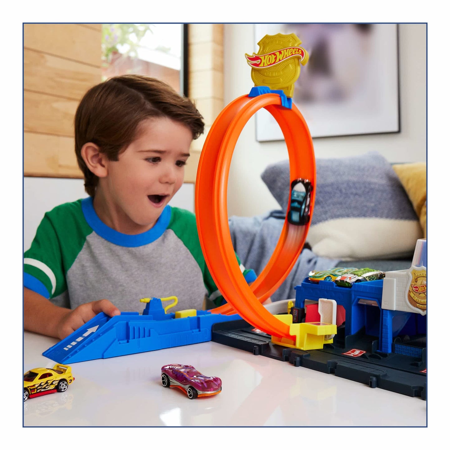 A boy excitedly watches a Hot Wheels City Super Police Station Tracks and Playset with an orange loop track and police station, as a car zooms through the loop