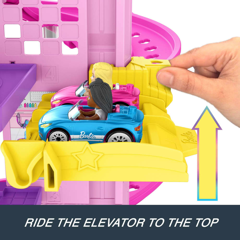 A hand lifts a yellow elevator carrying two Barbie dolls in cars up a pink multi-story playset