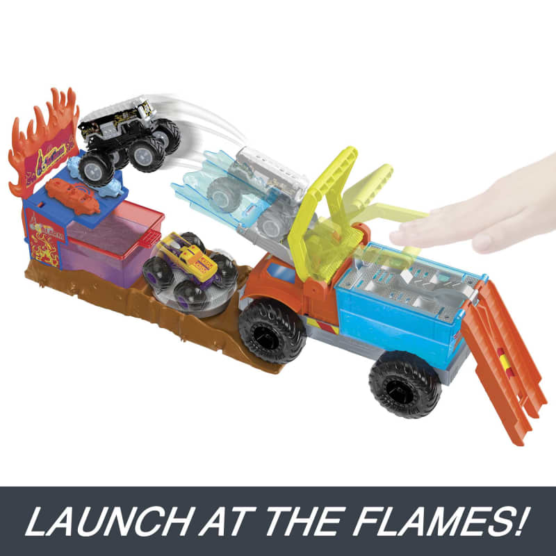Black toy monster truck jumps from a large orange and blue truck playset, flying over a yellow car towards fiery targets