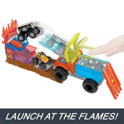 Black toy monster truck jumps from a large orange and blue truck playset, flying over a yellow car towards fiery targets