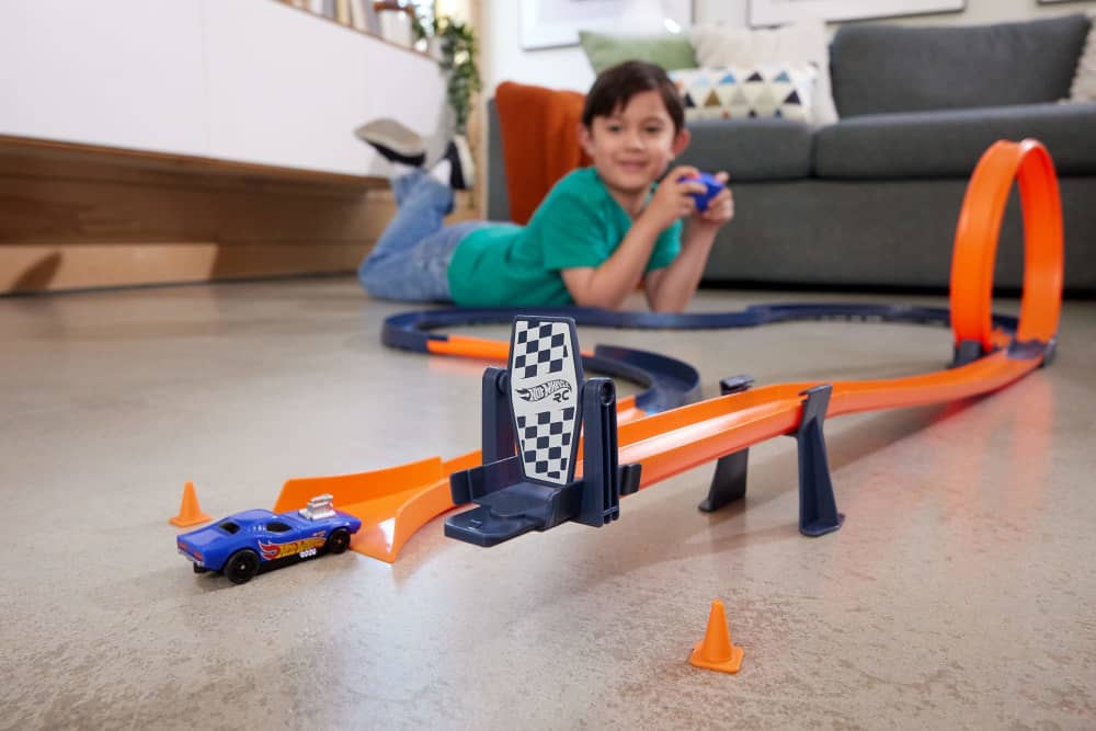 Happy child lies on floor, controlling a blue Hot Wheels RC car on an orange track with a loop and finish line
