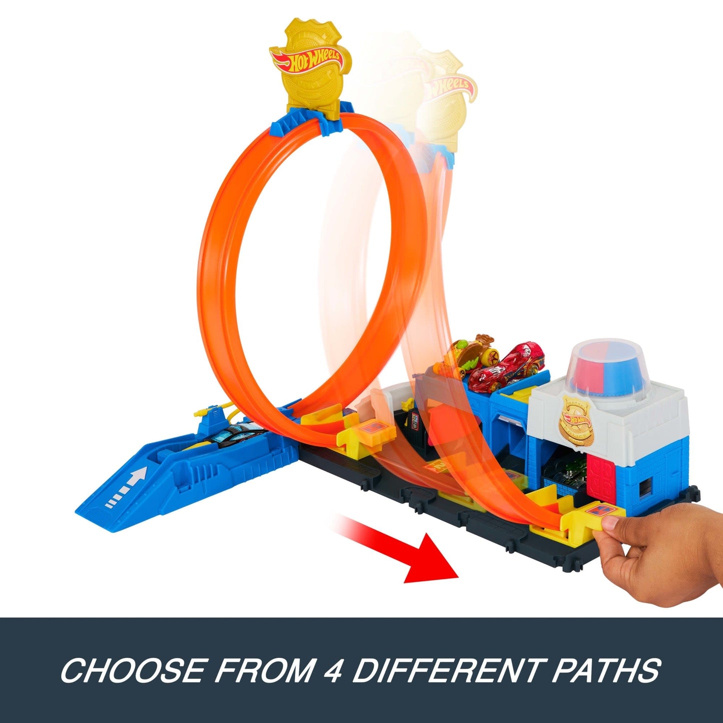 A police station playset with an orange loop track, a blue launcher, toy cars, and a hand adjusting a yellow track switch