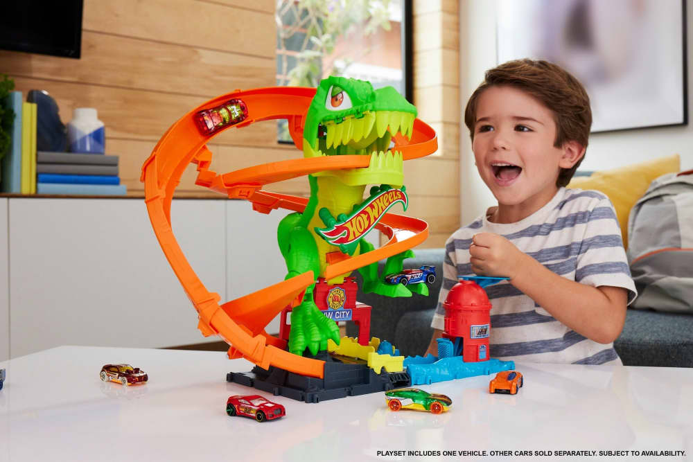 Smiling boy watches a Hot Wheels dinosaur playset with cars on orange tracks