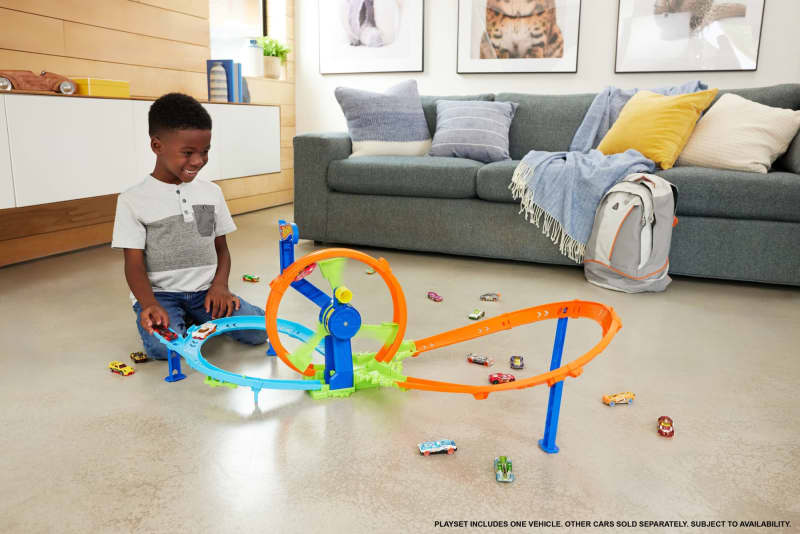 Smiling boy playing with a colorful toy car track, featuring a loop and launcher, surrounded by scattered toy cars on a living room floor