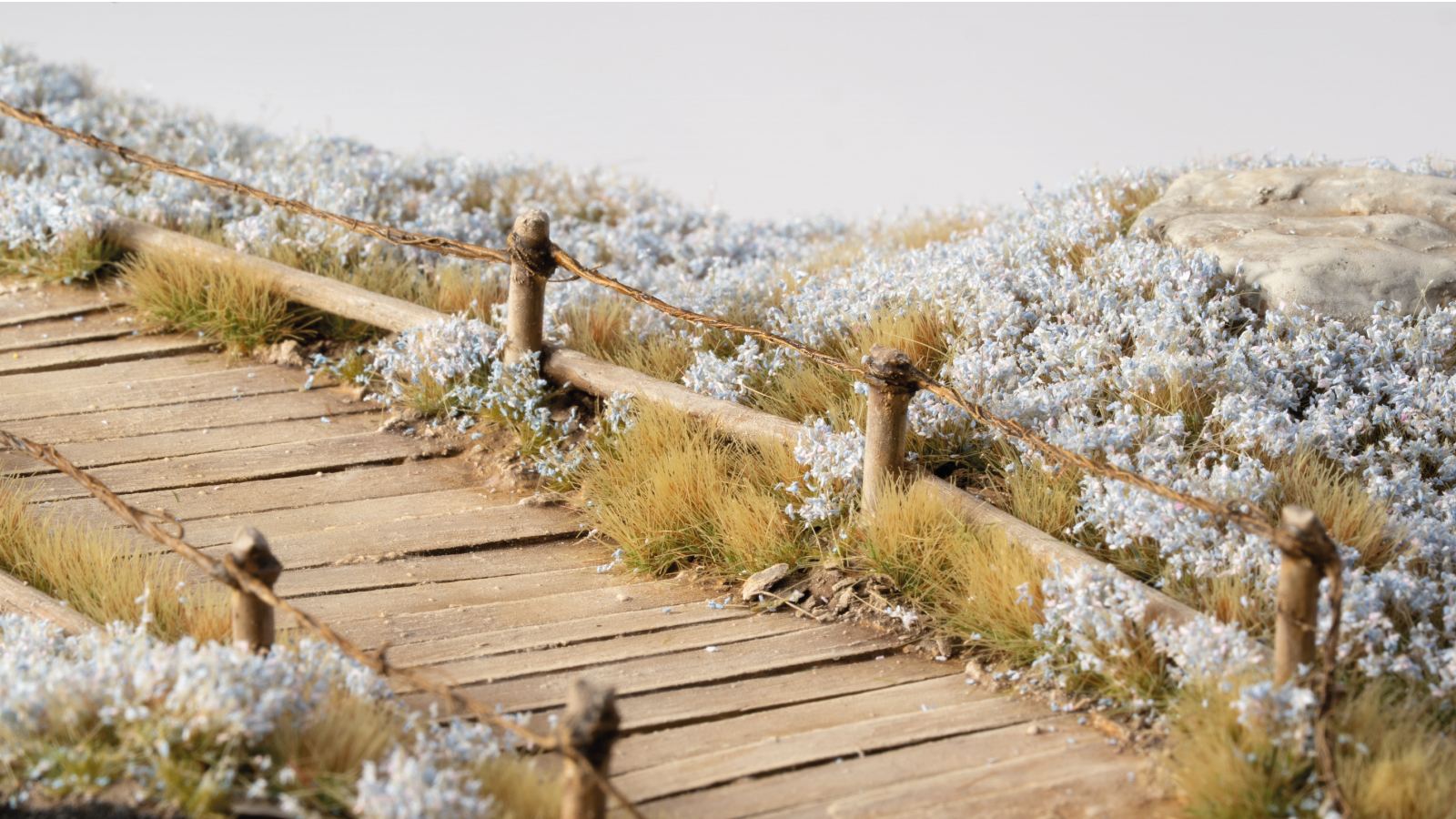 Detailed miniature wooden path with rustic wire fencing, surrounded by dense white flowers and dry grass