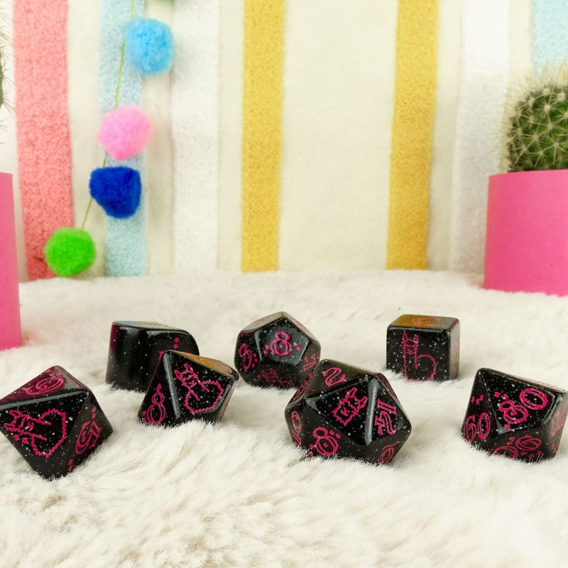 A set of seven black dice with pink numbers and cat designs scattered on a white fluffy surface.
