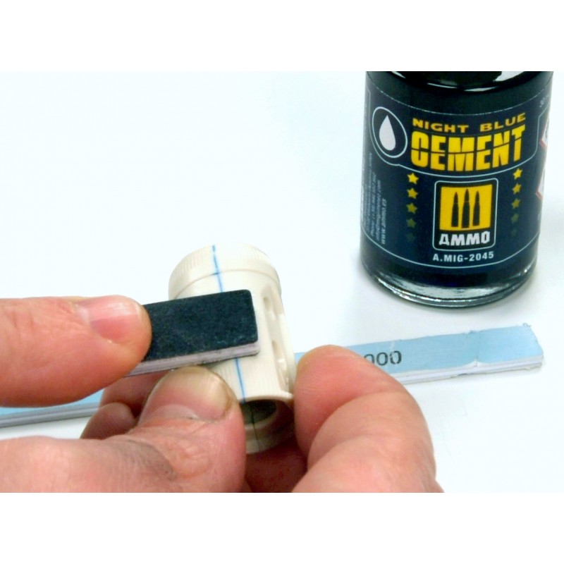 A hand sands a small white model part with a sanding block and an "AMMO Night Blue Cement" bottle is in the background