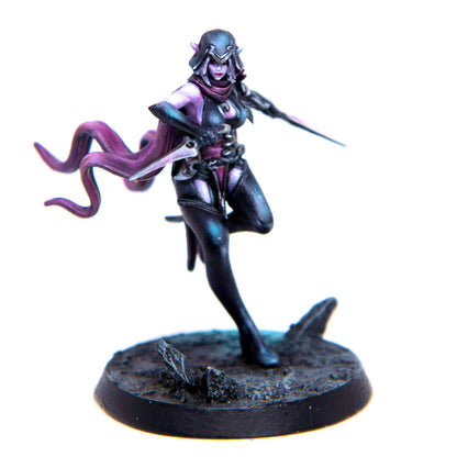 A miniature female dark elf with purple skin, hooded cloak and dark suit, lunging with two daggers on a rocky base.