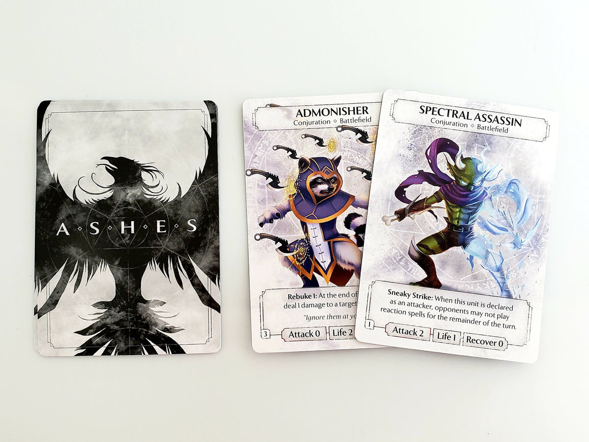 Ashes Reborn The Spirits of Memoria playing cards featuring the black and white Ashes card and two vibrant conjuration unit cards Admonisher and Spectral Assassin on a white surface