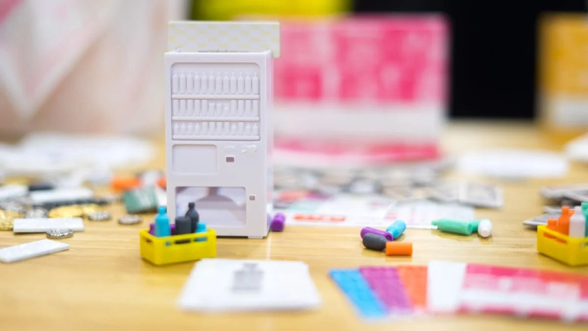 A miniature Tokyo Jidohanbaiki vending machine on a wooden table, surrounded by tiny colourful bottles in crates, cards, and small tokens