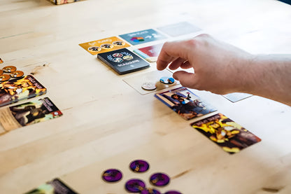 A hand moves a round token on a game card, surrounded by other game components on a light wooden table
