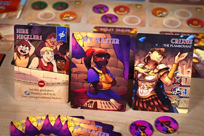 Three vibrant Gladius board game character cards: "Hire Hecklers" (a crowd), "Spy Master" shushing, and "Crixus the Flamboyant" performing