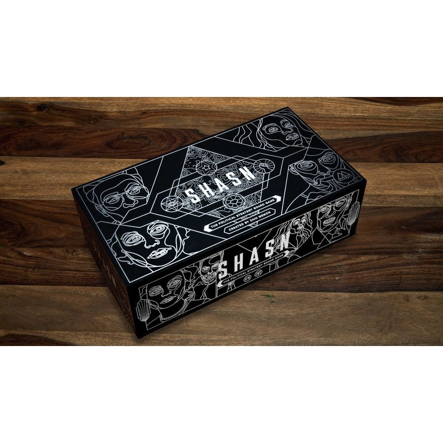 Black "SHASH" political strategy board game box with intricate silver line art depicting faces and geometric patterns, resting on a dark wooden table