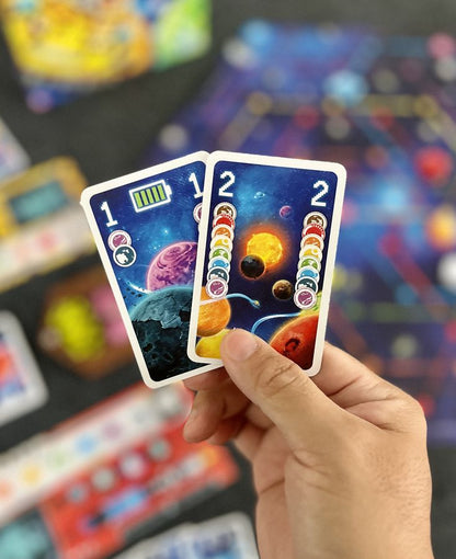 Hand holding two space-themed board game cards featuring planets, stars, resource icons, and numbers (1, 2)