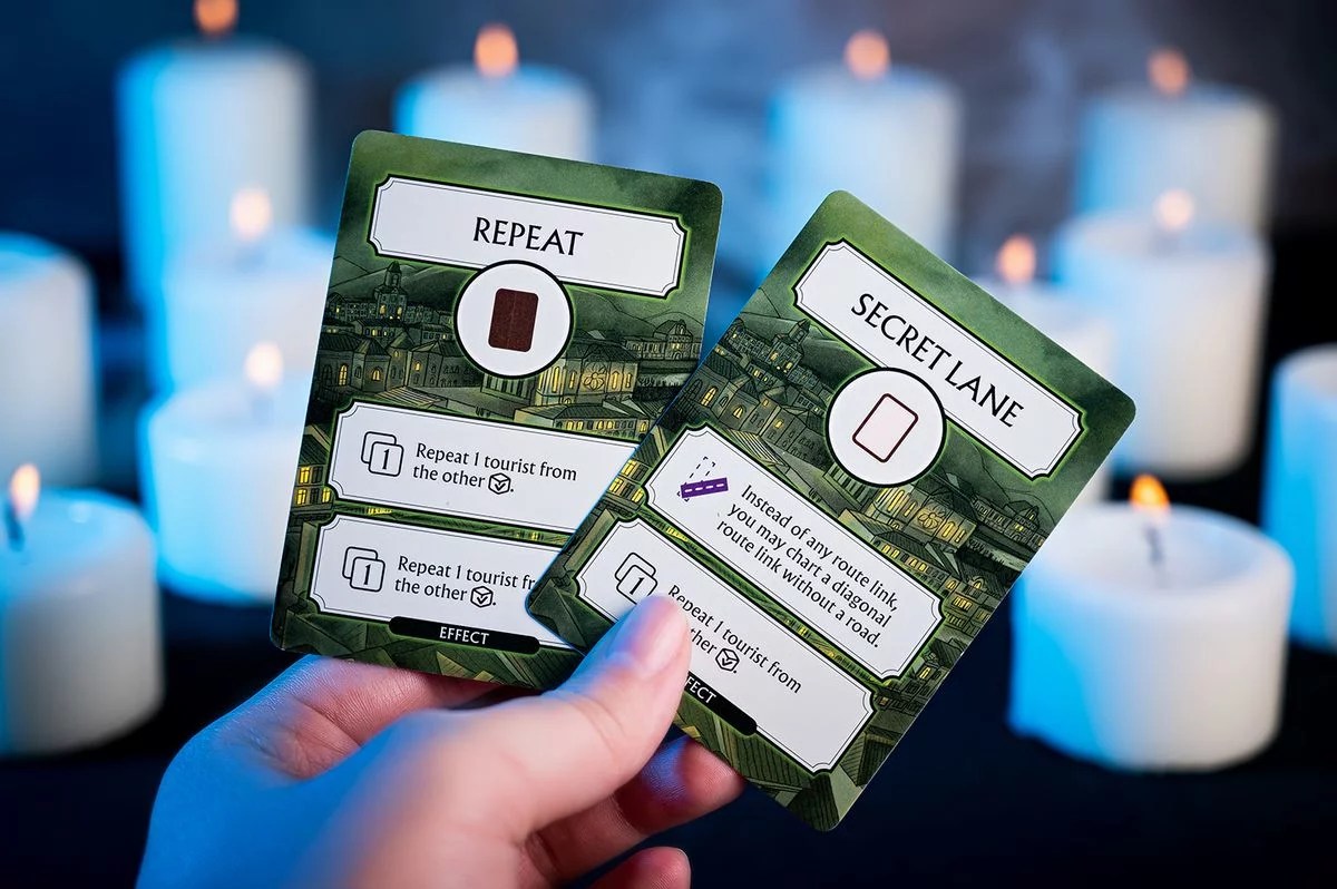 Hand holding two "Repeat" and "Secret Lane" green game cards, illuminated by many white flickering candles in the dark