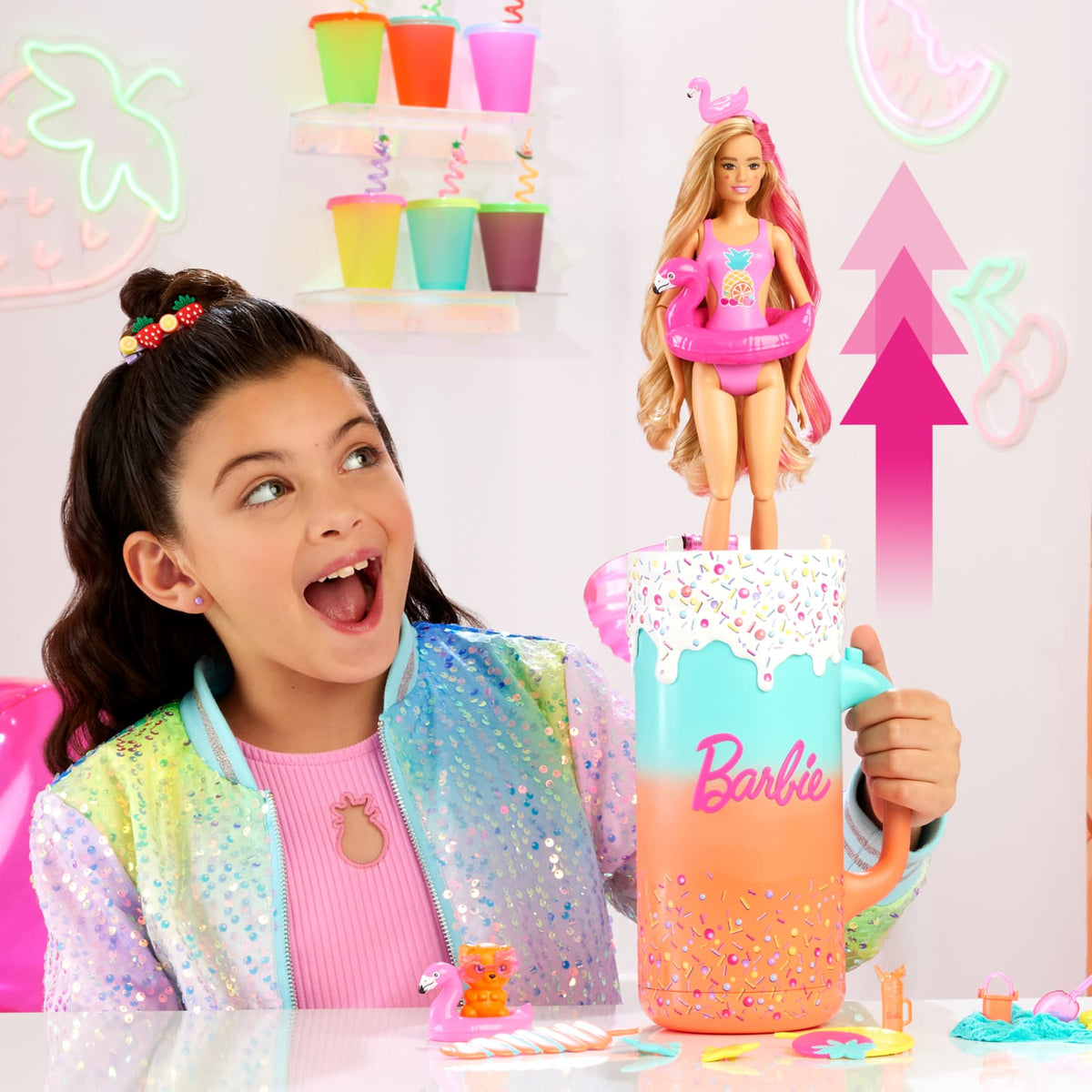 An excited girl in a sequin jacket looks at a Barbie doll in a swimsuit, emerging from a large, sprinkle-decorated Barbie mug