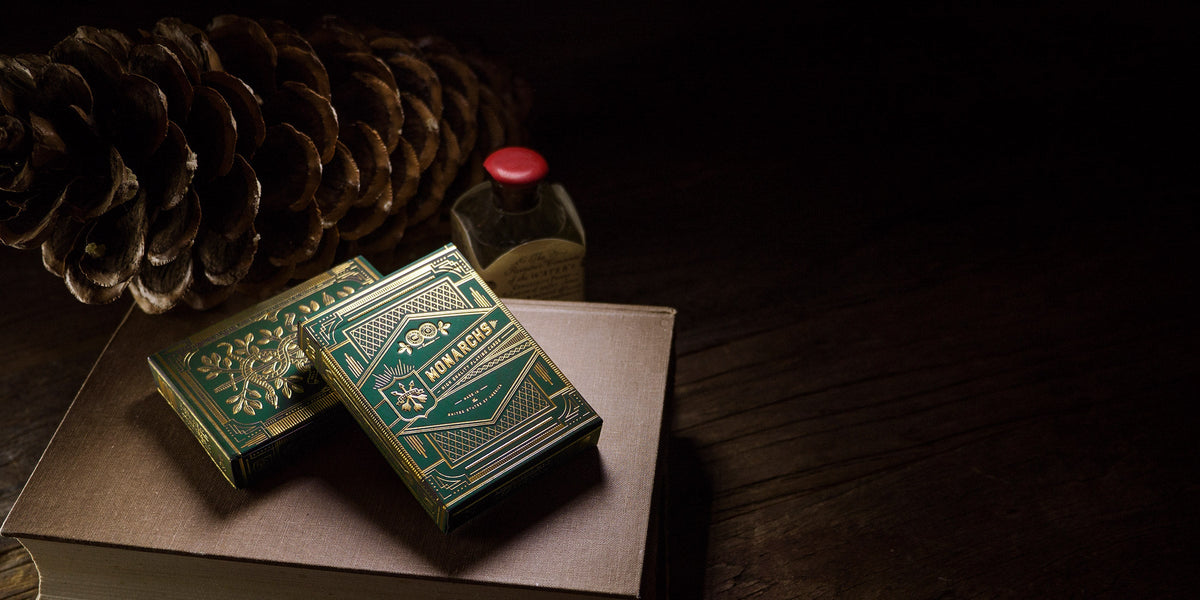 Two ornate green and gold Monarchs playing card decks rest on a brown book, with a pine cone and bottle in dim light