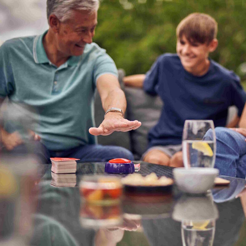 Smiling man and boy playing a game outdoors. The man&#39;s hand hovers over a red buzzer on a table with cards and drinks