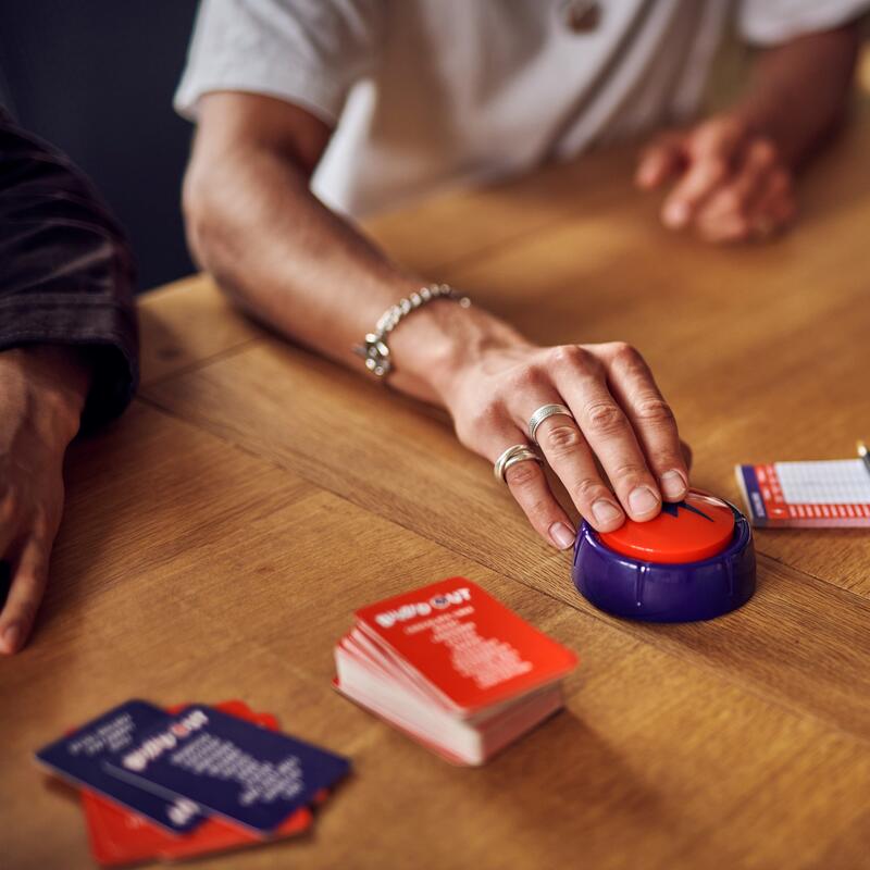 A hand presses a red game buzzer button on a wooden table with stacks of game cards.