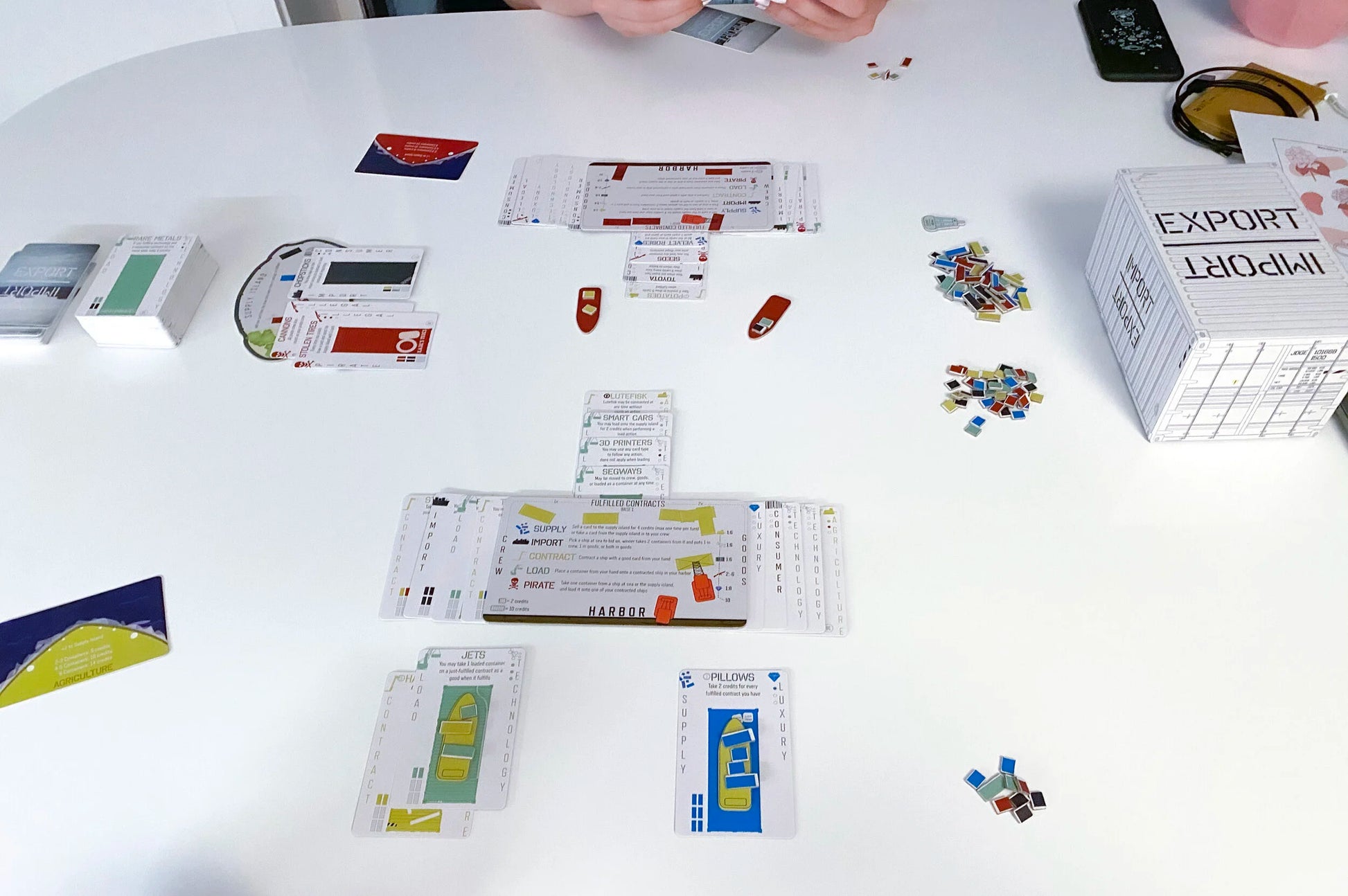 A white table displays an active board game with cards, colourful tokens, and a prominent EXPORT IMPORT shipping container box