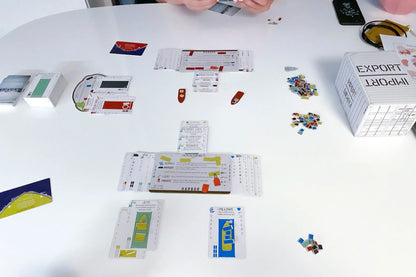 A white table displays an active board game with cards, colourful tokens, and a prominent EXPORT IMPORT shipping container box