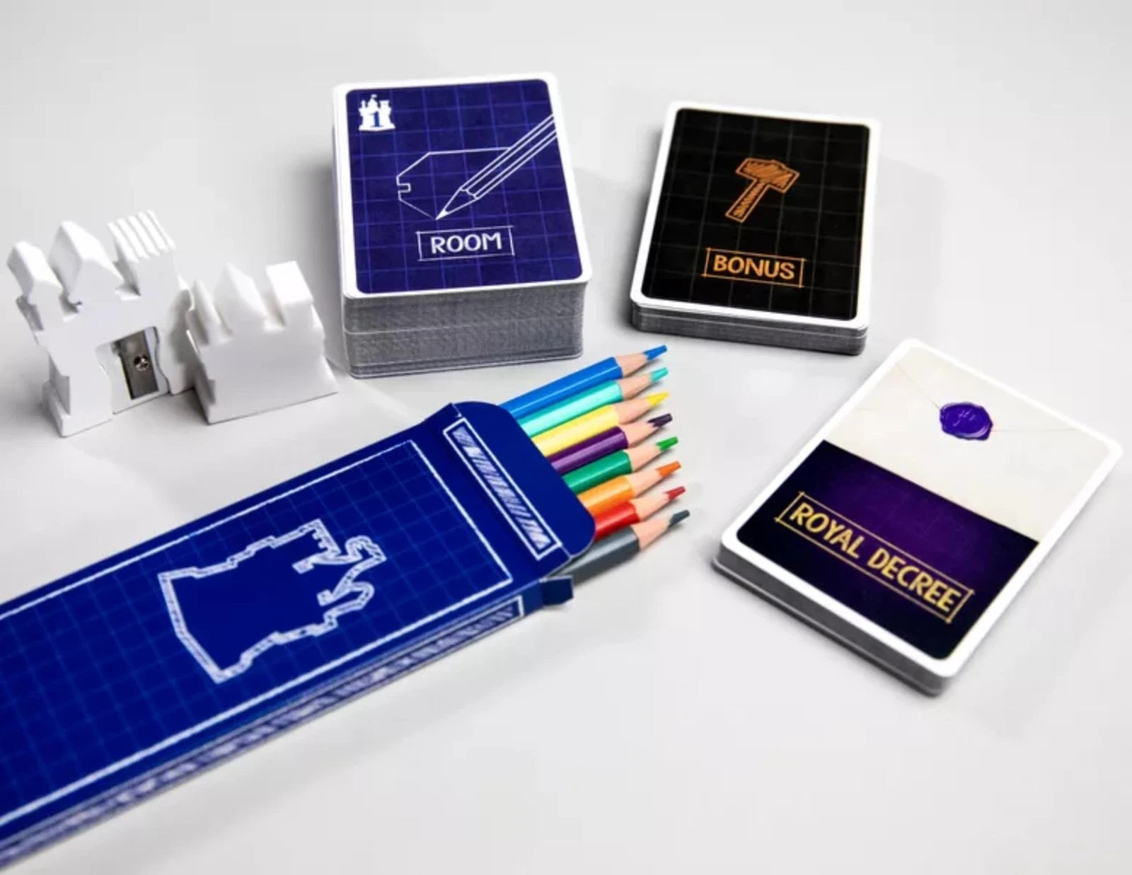 Castle-themed game components: stacks of cards, a white castle-shaped sharpener, and a blueprint pencil case with colored pencils