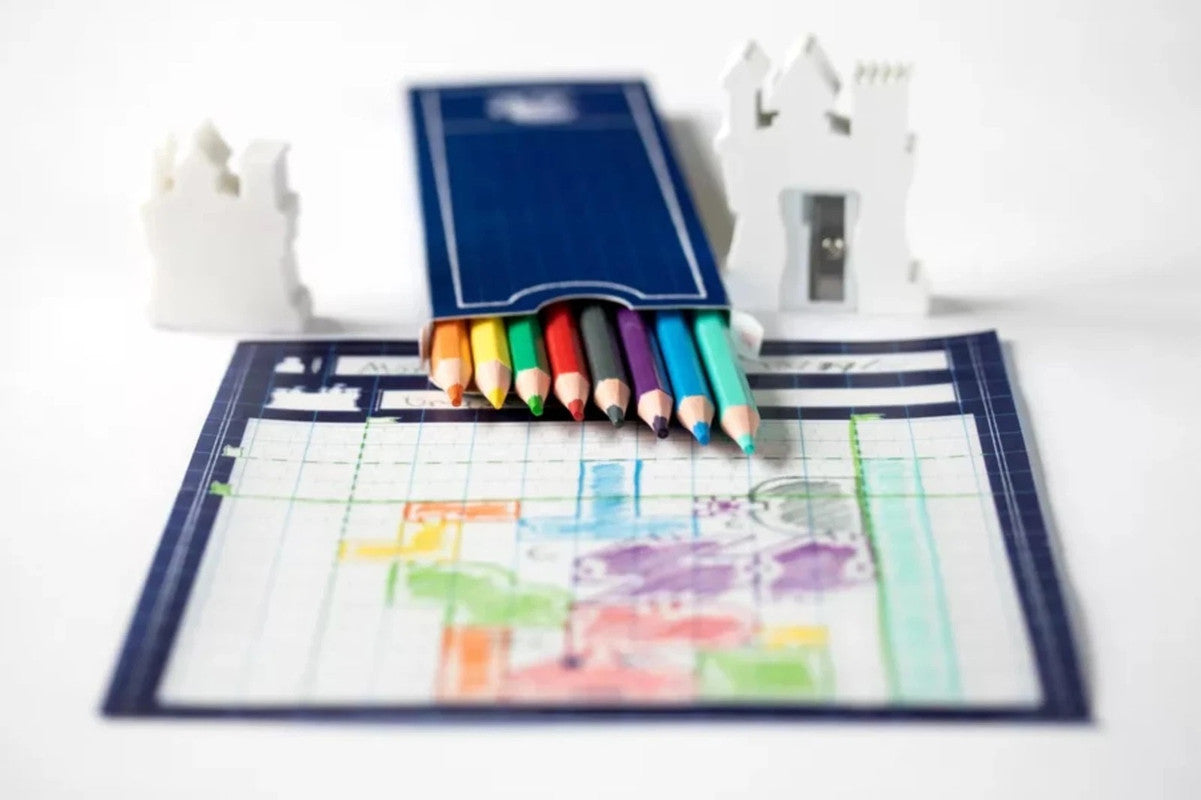 A set of colorful pencils in a blue box rests on a grid drawing with colored shapes, next to a castle sharpener