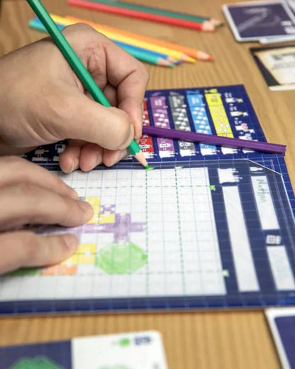 A hand uses a green pencil to fill in a grid on a board game sheet, surrounded by other pencils and cards