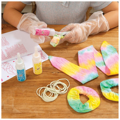 Gloved hands applying dye from a bottle to fabric, surrounded by tie-dye supplies (dyes, rubber bands) and finished colourful items