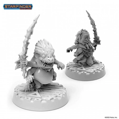 Two unpainted Starfinder Skittermander miniatures of a grinning, squat reptilian creature holding a jagged sword, seen front and back